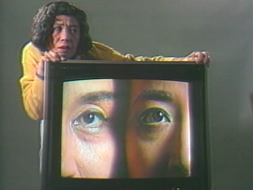 Tv screen with two faces side by side only showing half of each face. Artist behind the tv grasping both side of the screen with their face resting on left hand