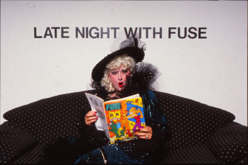 Artist Tanya Mars reading FUSE magazine, with a surprised face. Behind her reads letter in black "LATE NIGHT WITH FUSE"