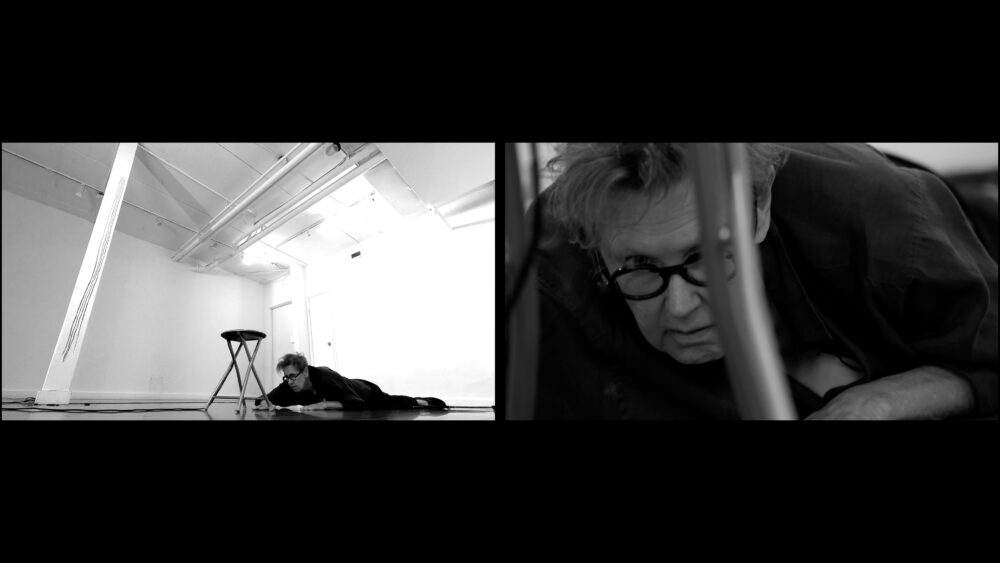 Two black and white images are side by side in a split screen. In the image on the left, we look up towards the ceiling of a big white room with a white column on the left. A woman in dark clothing is leaning over a metal stool. She is on her knees with one arm reaching to the floor and the other arm folded under her chest. The end of a walking stick is visible on the floor to the right. In the image on the right, a woman with short hair and glasses and wearing a dark shirt is lying awkwardly on the top of a metal stool. She has one arm folded under her chest and the other is reaching down. Her head is leaning over the edge of the stool facing down. Her body fills most of the picture frame.